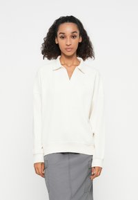 Suéter crema con cuello y escote en V. Hecho de una tela suave, presenta hombros caídos y puños acanalados. Llevado sobre una falda cargo gris.