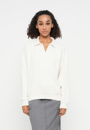 Suéter crema con cuello y escote en V. Hecho de una tela suave, presenta hombros caídos y puños acanalados. Llevado sobre una falda cargo gris.