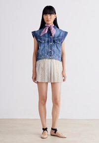Žena stojí oblečená v bezrukávom džínovom veste, plisovanej béžovej sukni, fialovej bodkovanej šatke na krku a béžových balerínach s čiernymi členkovými remienkami.
