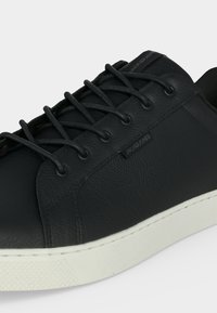 Baskets basses noires avec lacets ronds noirs et semelle en caoutchouc blanche, dessus en cuir texturé et petite étiquette logo sur le côté.
