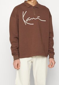 Maglione felpa marrone con colletto e mezza zip, caratterizzato da un logo ricamato bianco sul petto. Tessuto morbido e vestibilità comoda.