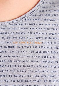 Šedé tričko z látky s opakujícími se textovými frázemi v námořnické modři a oranžové barvě. Design obsahuje kulatý výstřih a krátké rukávy.