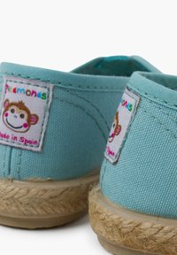Espadrile verzi din pânză cu tălpi din sfoară de iută. Include o etichetă cusută cu un grafic cu maimuță și textul "Fabricat în Spania".