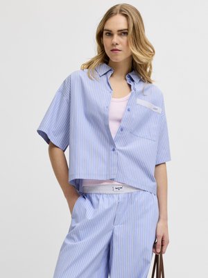 Młoda kobieta w jasnoniebieskiej i białej koszuli z pionowymi paskami na krótki rękaw oraz dopasowanych spodniach, pod spodem biała koszulka na ramiączkach, trzyma brązową torbę.