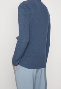 Blå stickad tröja med ribbad textur och långa ärmar, tillsammans med ljusblå byxor. Fickor synliga på sidan.