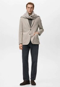 Beiger Blazer aus Wollmischung mit zwei Fronttaschen, grauer Pullover um den Hals gelegt, dunkle Jeanshose und braune Schuhe.