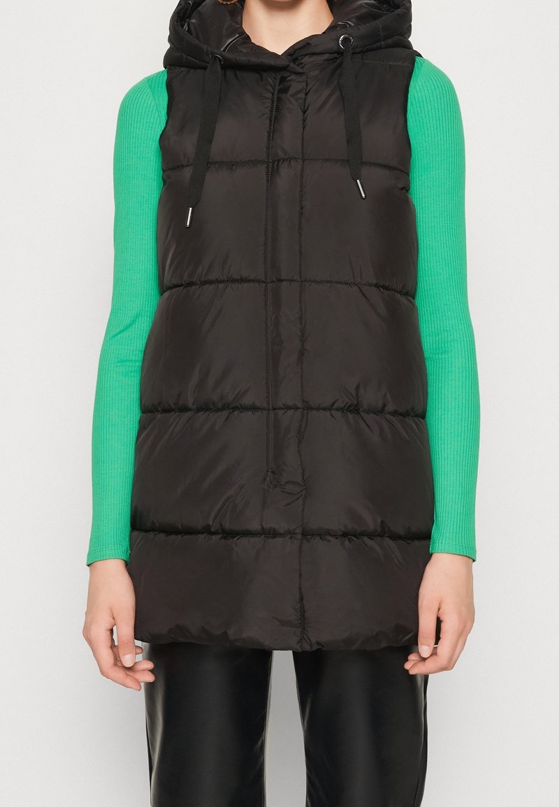 Schwarze Steppweste mit Kapuze, ausgestattet mit einem gesteppten Design und Kordelzügen. Kombiniert mit einem langen, gerippten grünen Oberteil.