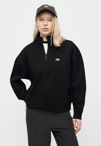 Νέα γυναίκα με ίσια ξανθά μαλλιά φοράει μαύρο φούτερ με φερμουάρ και καπέλο που ταιριάζουν, στέκεται μπροστά σε απλό φωτεινό φόντο.