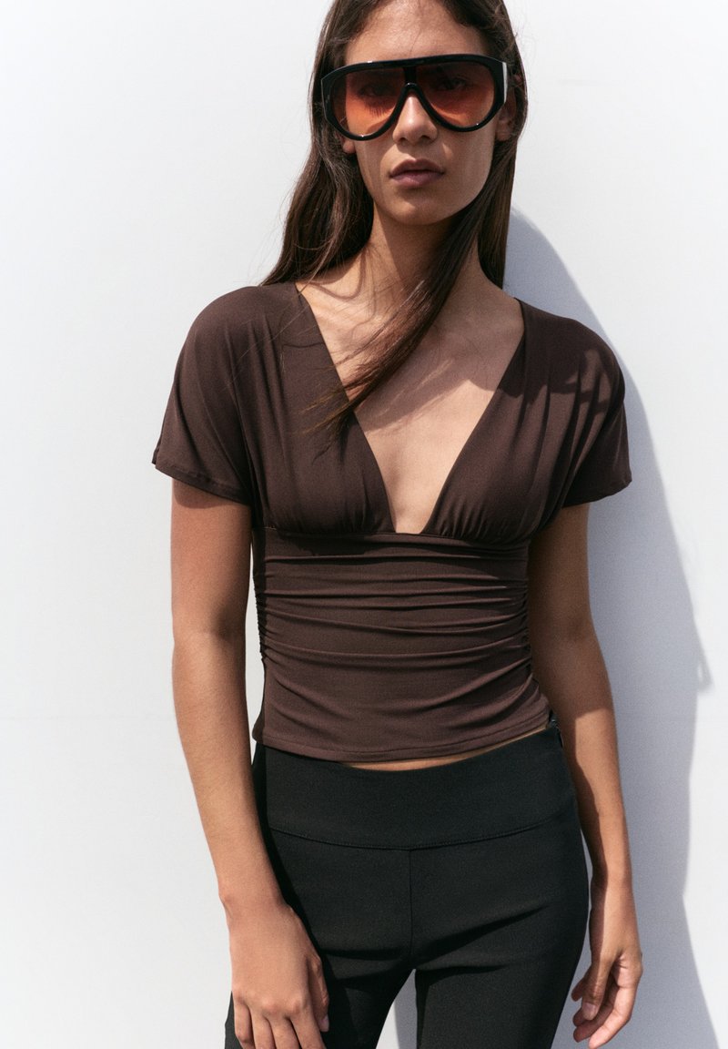 Jeune femme portant de grandes lunettes de soleil marron, un top marron près du corps avec un profond décolleté en V et un pantalon noir, debout contre un mur blanc.