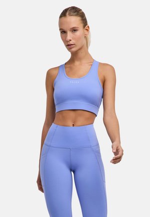 Frau trägt einen hellblauen Sport-BH und hochtaillierte Leggings und steht vor einem einfarbigen hellen Hintergrund.