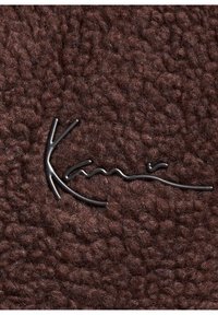 Tessuto marrone con una texture morbida e pelosa, caratterizzato da un logo in metallo scuro in uno stile di scrittura fluente.