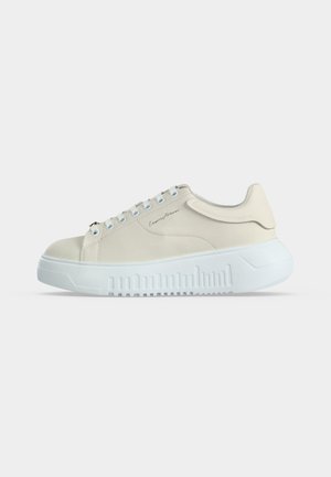 Nízký béžový teniska s bílou tlustou podrážkou, zavazováním vpředu a jemným logem ve stylu písma na boku, zobrazena z vnější strany.