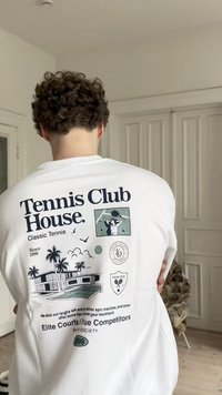 Pessoa com cabelo encaracolado vestindo uma camisa branca com gráficos e texto temáticos de ténis, de pé dentro de uma sala com portas brancas.