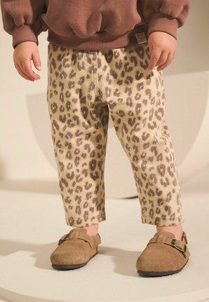 Maluch ubrany w beżowe spodnie w panterkę, brązowy sweter i brązowe buty na klamrę stoi na jasnej powierzchni.
