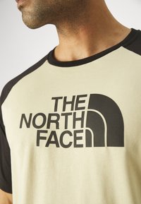 Kortærmet t-shirt i beige med sorte raglanærmer, der har et fremtrædende sort logo "THE NORTH FACE" på forsiden. Glat tekstur.
