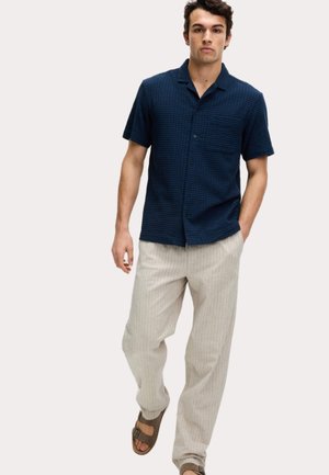 Navy short-sleeve shirt with a textured pattern, pocket on left chest, paired with light, striped, relaxed-fit trousers and brown sandals.