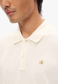 Πολυεστερικό μπλουζάκι σε ανοιχτό μπεζ χρώμα με υφή, διαθέτει κλασικό γιακά, τρία κουμπιά και ένα μικρό κεντημένο λογότυπο σε χρυσό στην περιοχή του στήθους.