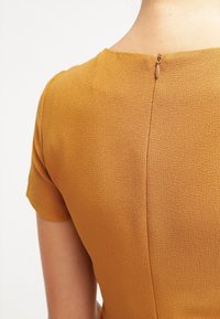 Kortärmad klänning i senapsgul tyg med en strukturerad yta. Har en diskret blixtlås bak och en hög halsringning.