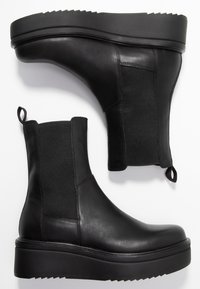Botas negras de tobillo con un upper de cuero suave, paneles laterales elásticos, lengüetas para tirar y una suela gruesa y estriada para mayor altura.