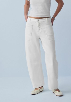 Femeie purtând pantaloni albi cu talie înaltă și croială dreaptă, cu buzunare aplicate, top alb cu mâneci scurte și pantofi crem cu talpă joasă pe fundal albastru deschis.