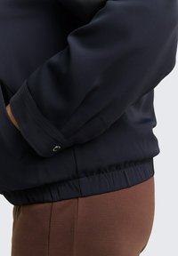 Veste bleu marine avec une texture lisse, ceinture élastique et poignets boutonnés. Gros plan montrant la manche et la zone des hanches pour les détails d'ajustement.