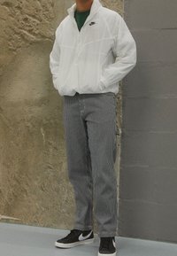 Vit nylonjacka med dragkedja och svart logotyp, i kombination med grå- och vitrandiga byxor och svarta sneakers med vita accentuerade detaljer.