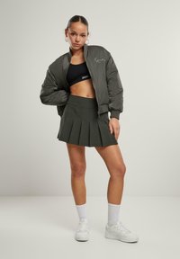 Olivgrüner Bomberjacke mit gerippten Bündchen, schwarzem Crop-Top und plissierter, dunkelgrüner Minirock. Weiße Sneakers und Crew-Socken vervollständigen das Outfit.