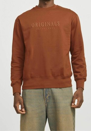 Mężczyzna noszący bluzę w kolorze rdzy z wypukłym napisem „ORIGINALS JACK & JONES” na piersi, w połączeniu z jasnoniebieskimi dżinsami.