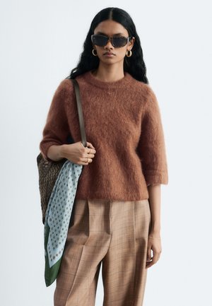 Femme portant un pull brun duveteux, un pantalon à carreaux, des lunettes de soleil écaille de tortue, des boucles d'oreilles créoles en or, portant un sac tissé avec une écharpe à motifs.