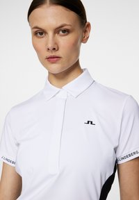 Vit pikétröja med krage, med en svart logotyp på bröstet och svarta detaljer på sidorna och ärmarna, tillverkad av slät tyg.