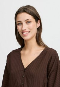 Brun texturerad blus med V-ringning, långa ärmar och knäppning. Tyget har ett subtilt randigt mönster f�ör extra detalj.