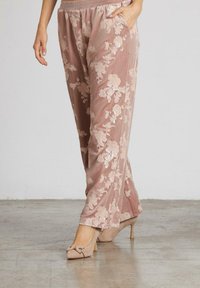 Donna che indossa pantaloni in velluto rosa a fiori con vita elastica e scarpe col tacco beige, in piedi su un pavimento in cemento.