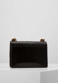 Černá obdélníková kabelka z kůže s raženým krokodýlím vzorem. Vyzdobena kovovým řetízkem na vrchu a má hladký, lesklý povrch.