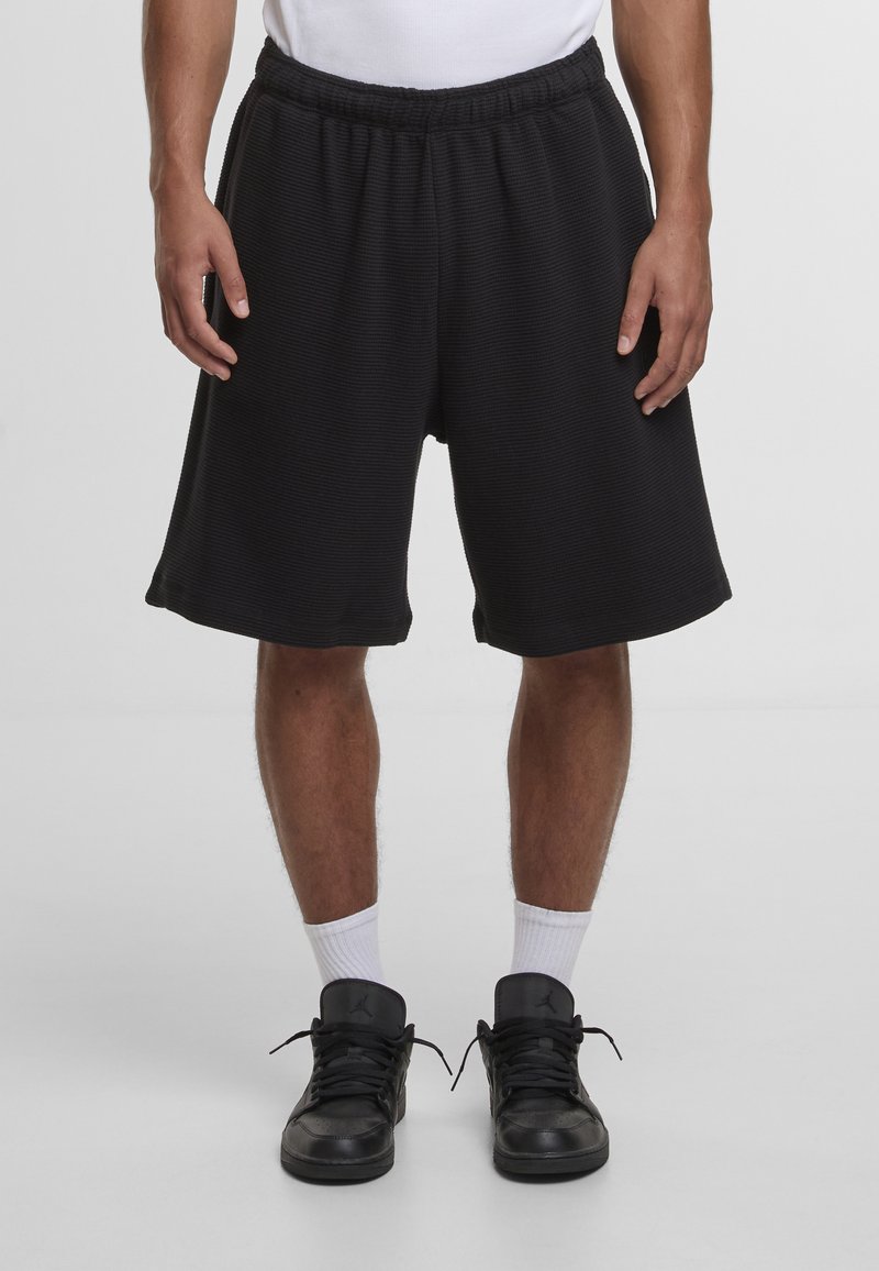 Shorts de sport noirs texturés avec une ceinture élastique, arrivant mi-cuisse, assortis à des baskets noires et des chaussettes blanches.