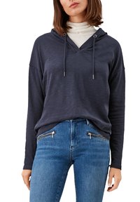 Tmavě modrá mikina s kapucí a výstřihem do V, s dlouhými rukávy a volným střihem, nošená přes bílý rolák, doplněná modrými džíny s kapsami na zip.