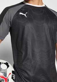 Camiseta de fútbol negra de Puma con mangas de malla gris, que presenta un sutil patrón de olas y un cuello blanco. El jugador sostiene un balón de fútbol.