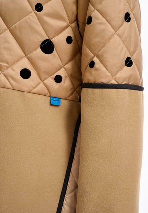 Prešívaná béžová bunda s čiernymi bodkami a modrou značkou. Má hladkú textúru a kontrastnú čiernu lemovku na spodnej časti.