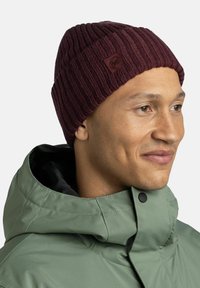 Burgundy ribbed knit beanie with a leather logo patch. Paired with a light green waterproof jacket featuring a high collar and snap closure.