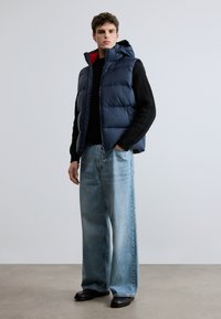 Colete acolchoado azul-marinho com interior vermelho, combinado com um suéter de malha preto e jeans de perna larga azul-claro, sobre um fundo neutro.