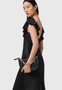 Zwarte satijnen jurk met gerimpelde mouwen en een lage rug, vastgehouden door een hand die een zwarte tas met een gouden kettingband draagt. Soepele textuur.