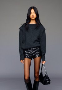 Šedý nadrozmerný sveter, čierne šortky so sequinkami, priehľadné pančuchy, čierne čižmy po kolená a čierna kabelka. Neutrálny pozadie.