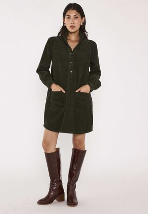 Donna che indossa un vestito di velluto a coste verde scuro con tasche frontali e stivali di pelle marroni al ginocchio, in piedi su uno sfondo uniforme.