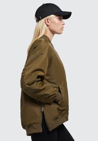 Olivovo zelená bomber bunda s rebrovaným pásom a manžetami, so zipsovou bočnou vreckom a čiernou čiapkou. Hladký, lesklý materiál, priliehavý strih.