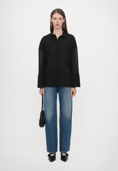 Zwarte doorzichtige blouse met lange mouwen en een kraag, gecombineerd met lichtblauwe rechtvallende spijkerbroeken en zwarte puntige ballerina's, vastgehouden met een kleine zwarte tas.