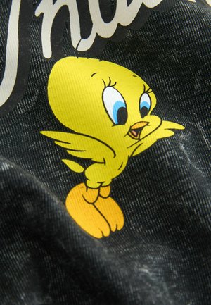 Gelber Cartoon-Vogel mit blauen Augen und offenem Schnabel, der auf orangefarbenen Füßen vor einem strukturierten schwarzen Stoffhintergrund steht.