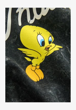 Sárga rajzfilm madár kék szemekkel és nyitott csőrrel, narancssárga lábakon állva, egy textúrált fekete szövet háttér előtt.