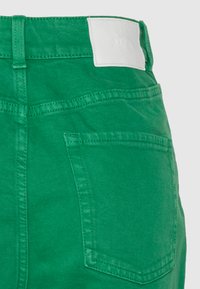 Shorts de ganga verdes com um bolso na parte de trás e um rótulo branco da marca na cintura. O tecido tem um acabamento liso, ligeiramente texturizado.