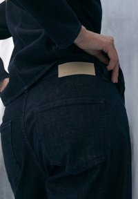 Jean en denim foncé avec une texture lisse, doté d'une pièce en cuir marron clair à la taille, avec les poches arrière visibles d'un côté.