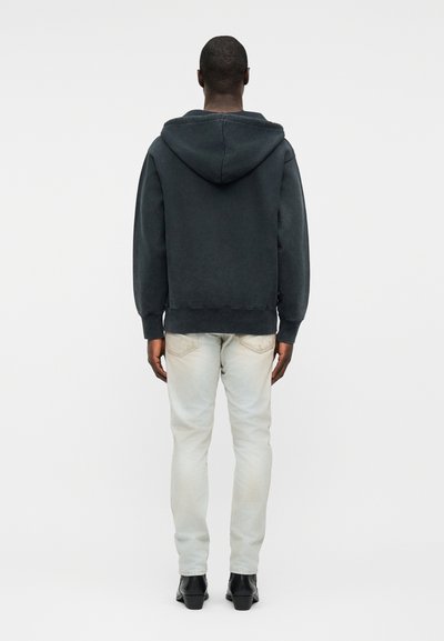 Homme debout de dos, portant un sweat à capuche noir, un jean clair et des chaussures noires sur un fond blanc uni.