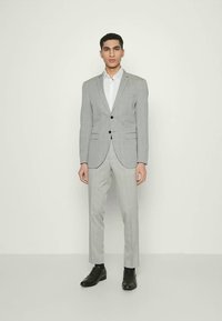 Jovem vestido com um fato xadrez cinza claro, uma camisa branca e sapatos pretos, de pé contra um fundo claro e uniforme.